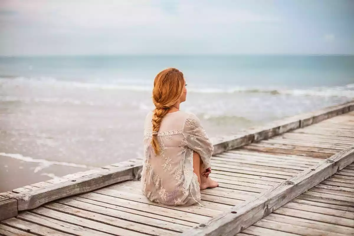 Una chica sentada agarrándose las piernas con las manos y mirando al mar