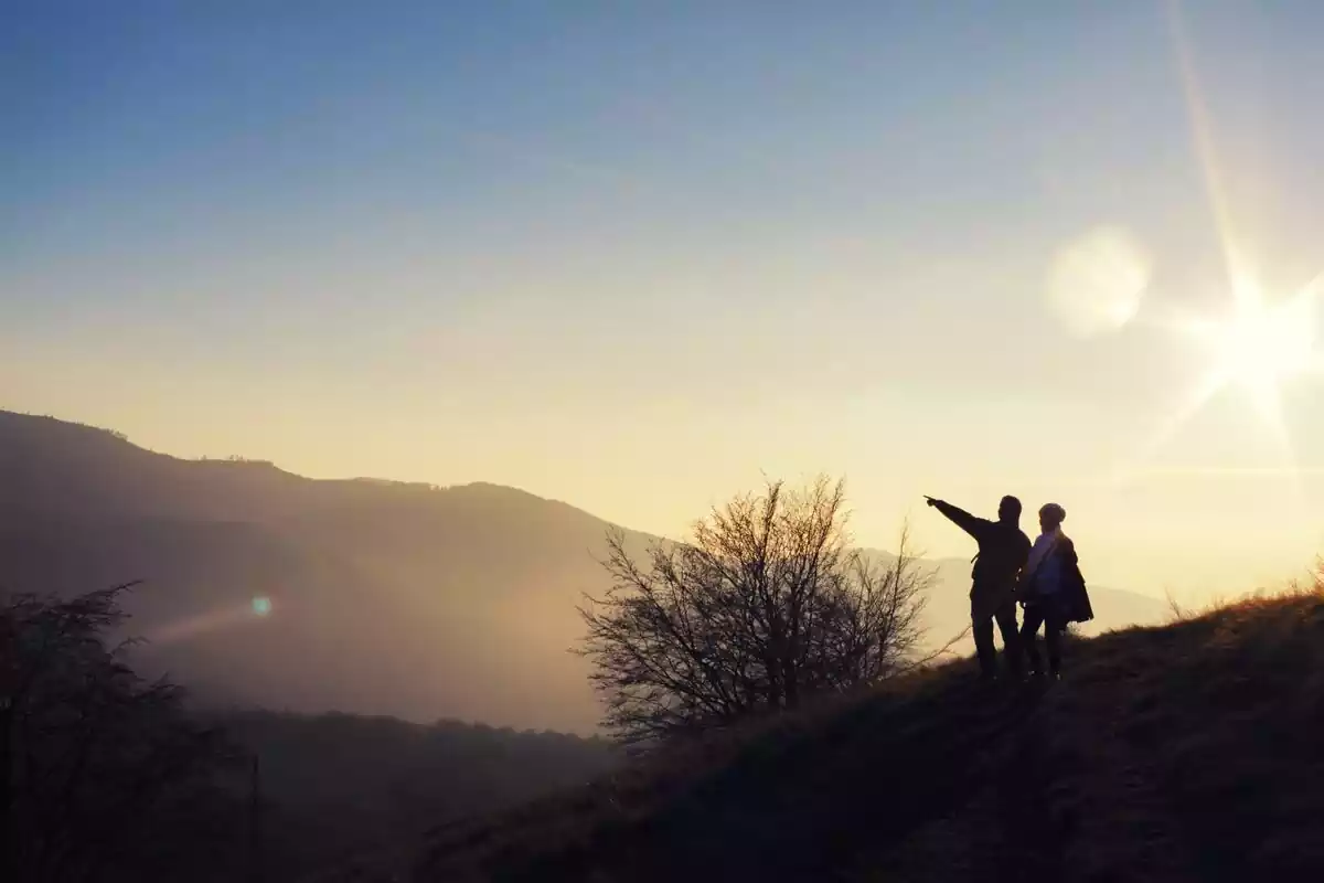 Dos personas situadas en una montaña señalando el horizonte con un atardecer de fondo