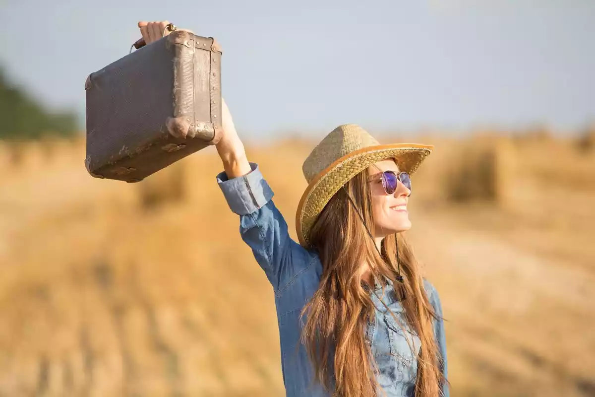 Una chica con una sonrisa mirando hacia el sol y una maleta en la mano