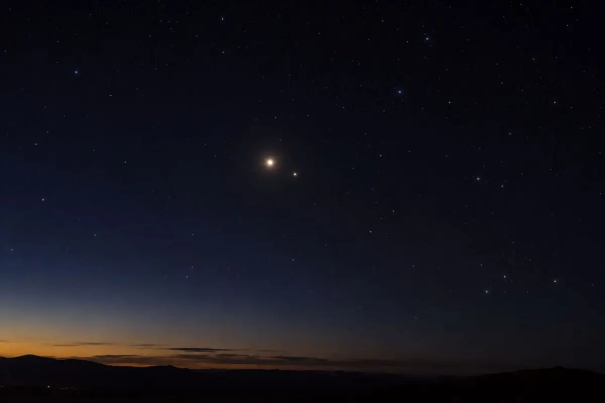 Cielo nocturno con dos planetas muy brillantes sobre el horizonte al anochecer y un paisaje de montañas oscuras.