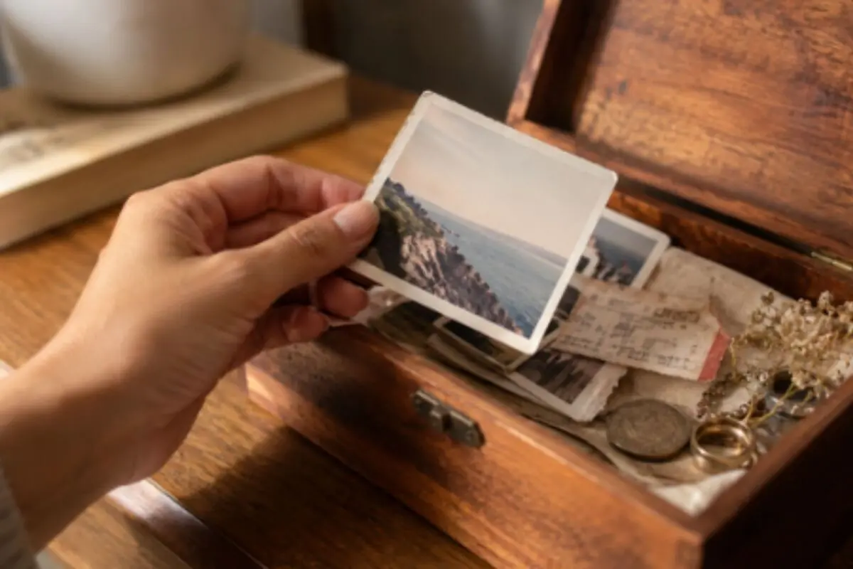 Mano sosteniendo una fotografía antigua sobre una caja de madera llena de recuerdos y objetos personales.