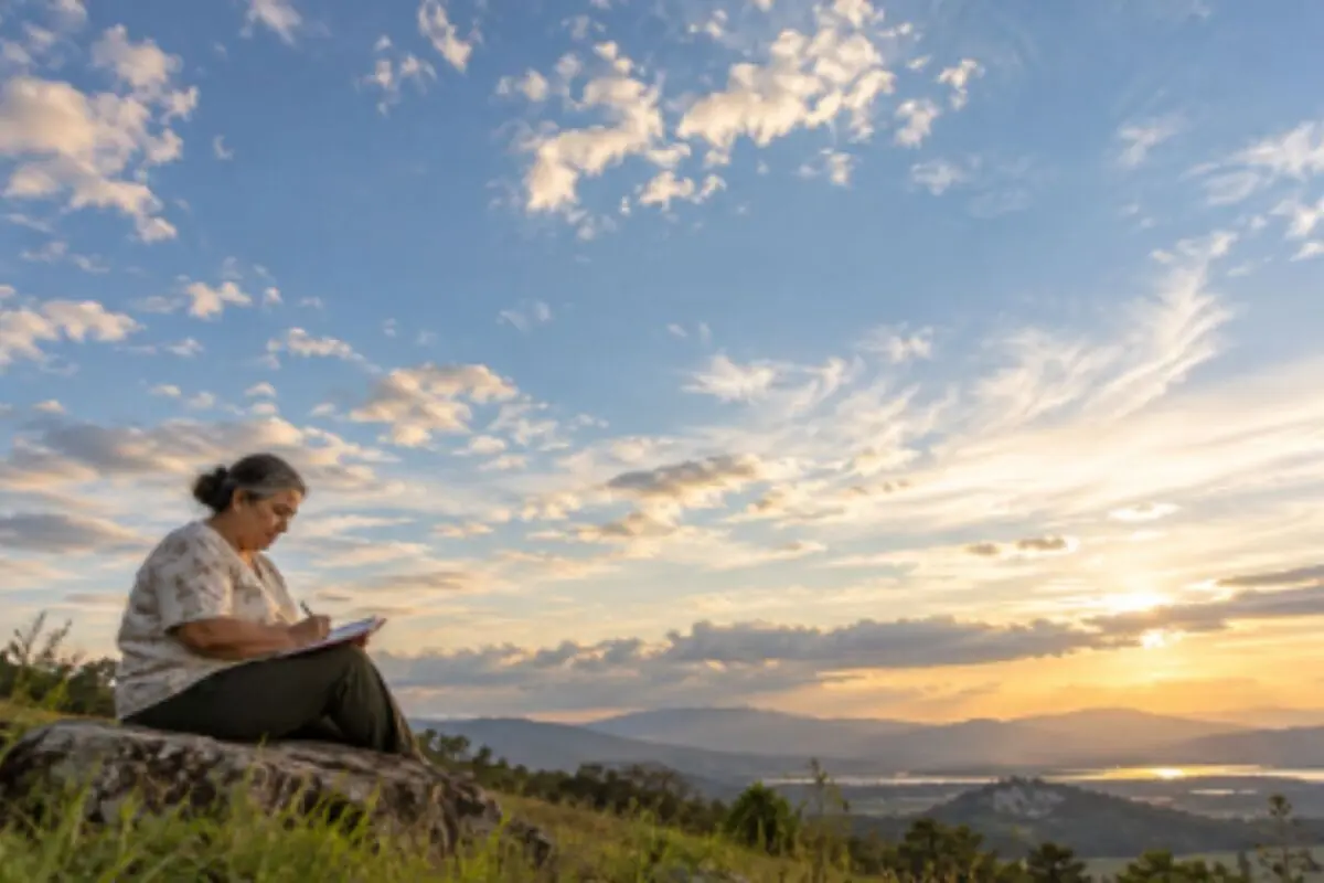 Mujer mayor sentada sobre una roca escribiendo en una libreta mientras contempla un atardecer en el campo con montañas y nubes en el horizonte. Mujer mayor sentada sobre una roca escribiendo en una libreta mientras contempla un atardecer en el campo con montañas y nubes en el horizonte.