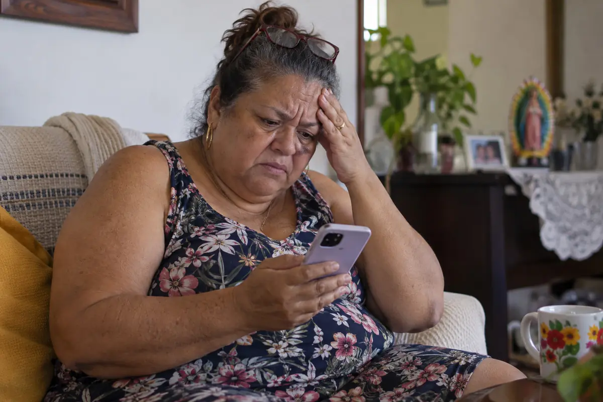 Mujer mayor con gesto de preocupación sentada en un sillón mientras mira la pantalla de su teléfono móvil en una sala de estar.