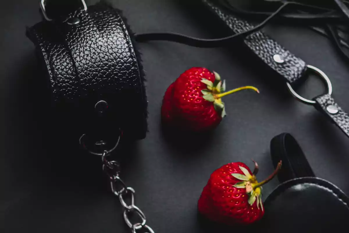 Dos fresas rojas junto a accesorios de cuero negro sobre un fondo oscuro.